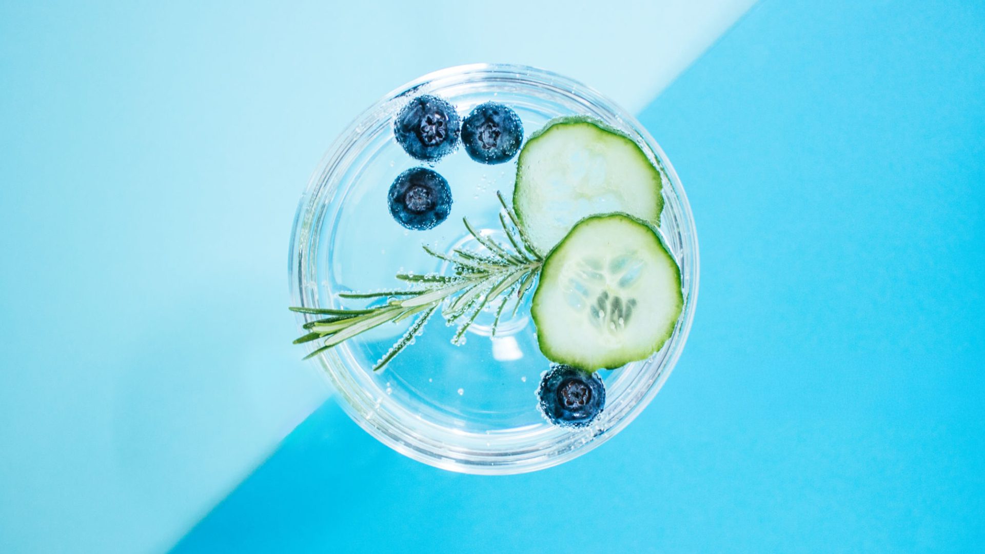 Overhead photo of a gin glass with cucumber slices, blueberries and rosemary on a light blue background.